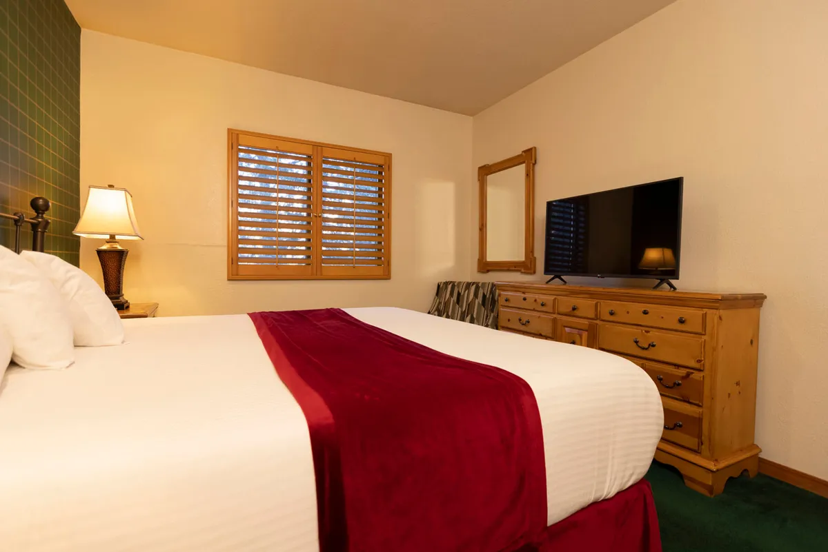A comfortable bedroom scene featuring a bed with a red runner, a window with classic shutters letting in gentle daylight, a wooden dresser with a lamp, and a TV. The space is styled for restful nights and peaceful mornings, emphasizing homeliness and privacy.