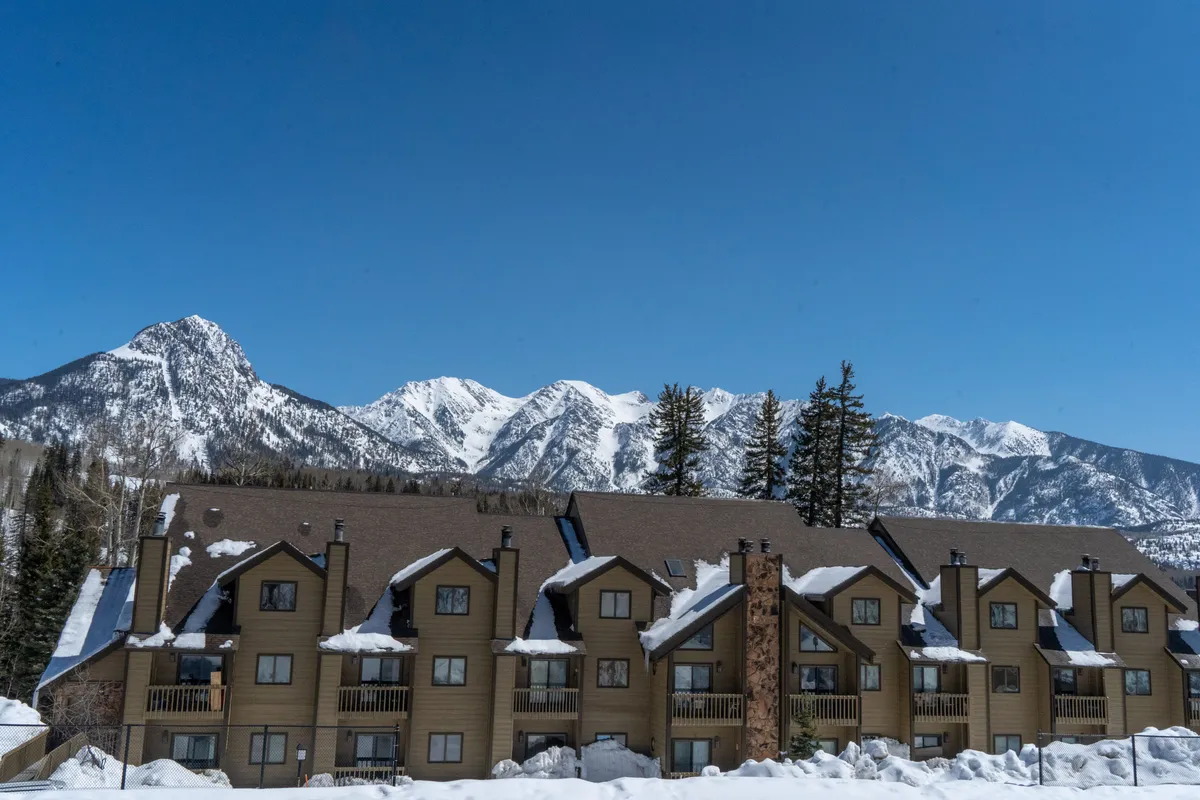 A striking exterior view of a mountain lodge with rustic wooden balconies and sturdy stone accents, set against a panorama of snow-covered mountains. The scene exudes a sense of comfort and adventure, hinting at cozy accommodations just steps from outdoor excitement.