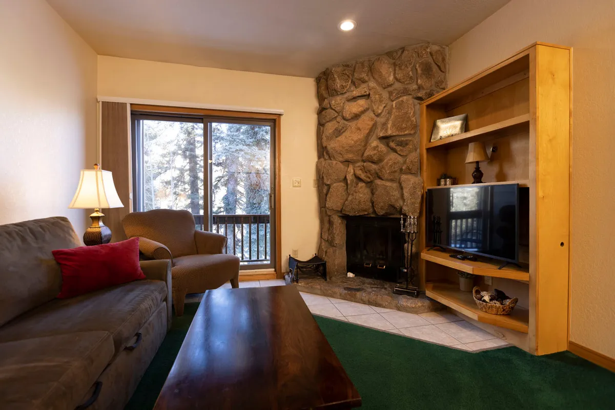 The image showcases a warm and inviting living area featuring a plush sofa, a rustic stone fireplace, a large window that allows abundant natural light, and a TV unit. Soft furnishings and cozy decor create a tranquil environment ideal for relaxing after a day in the mountains.