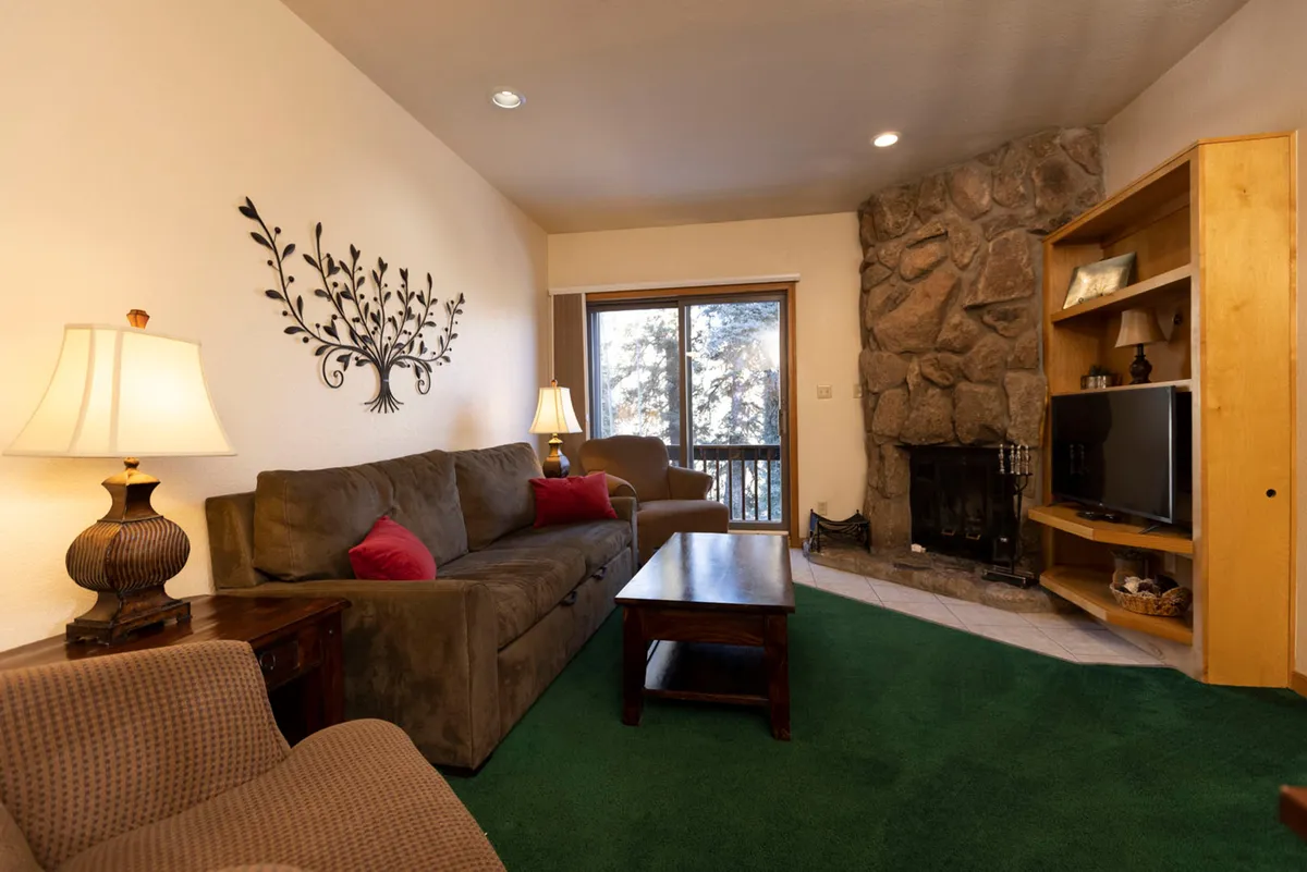 The image showcases a cozy living area in a Durango condo, featuring a plush sofa facing a crackling fireplace, a bookshelf filled with inviting reads, and large windows that offer natural light and scenic mountain views. The mood conveys warmth and relaxation, perfect for curling up after a day outdoors or sharing time with loved ones.