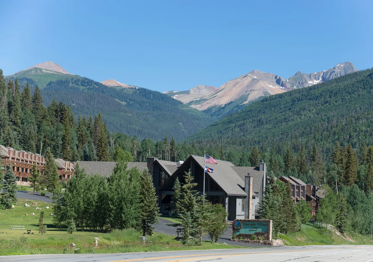 This image captures the exterior of Cascade Village, with the main lodge nestled amidst tall trees and stunning mountain scenery. The roadside sign and visible hiking trails suggest easy access to outdoor adventures, while the calm setting reflects the tranquil atmosphere of the resort. The image radiates a sense of seclusion and relaxation, with nature enveloping the property.