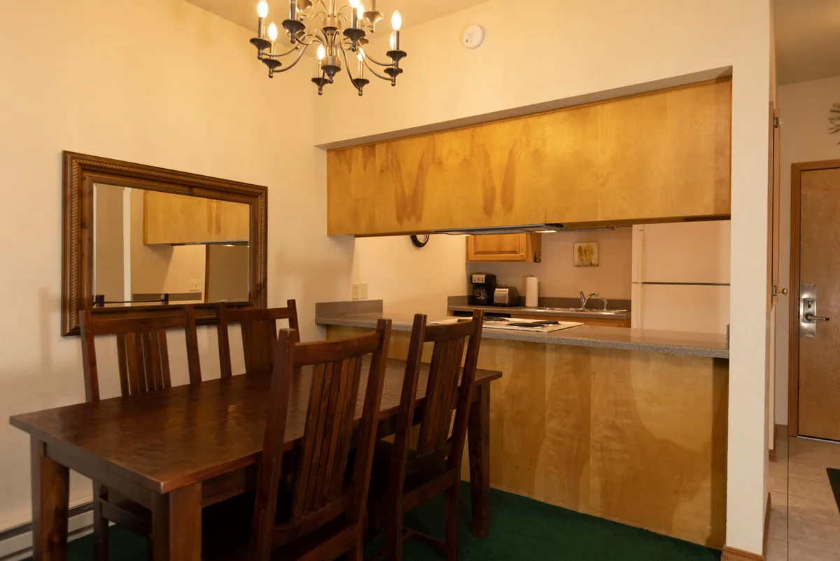 Featuring a wooden dining table under a chandelier, this image presents a cozy dining nook flowing into the kitchen area. The kitchen counter is visible, and the ambiance suggests intimacy and communal gatherings, ideal for relaxed family breakfasts or celebratory dinners.