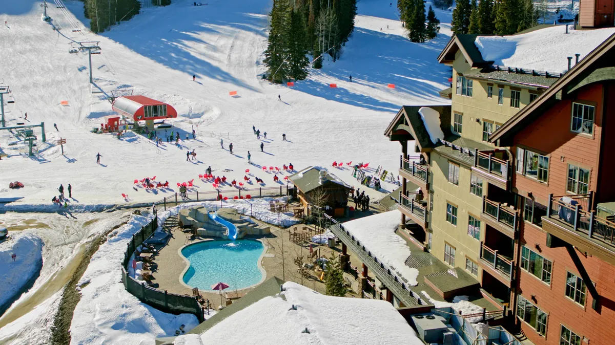 A lively scene showing a swimming pool adjacent to ski slopes and lodge buildings. Visible are skiers and snowboarders enjoying the area, with guests relaxing in the pool, capturing the blend of adventure and leisure at the heart of the property.