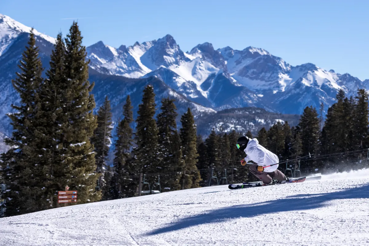 A skier is captured in motion as they glide down a snow-covered slope, framed by majestic, rugged mountains under a crisp blue sky. The energy and exhilaration of the ski experience are palpable, with untouched powder and sparkling snow creating a quintessential winter adventure scene.