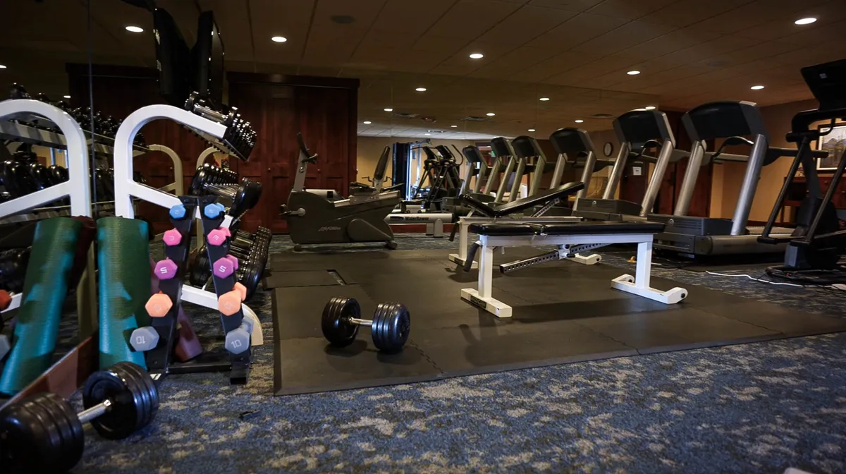 An interior fitness room featuring weightlifting equipment, treadmills, and dumbbells arranged in a clean, well-lit space. The environment is organized and clearly intended for guests seeking convenience and maintaining routines while traveling.