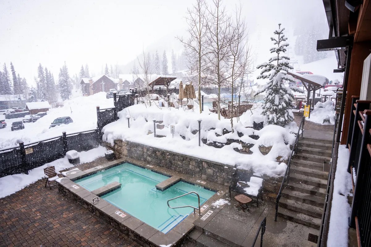 An inviting outdoor hot tub set against a snowy mountain backdrop, with steam rising and snow-covered trees all around. Steps lead down to the stone patio, offering space for gathering, while the serene landscape enhances the atmosphere of relaxation and indulgence.