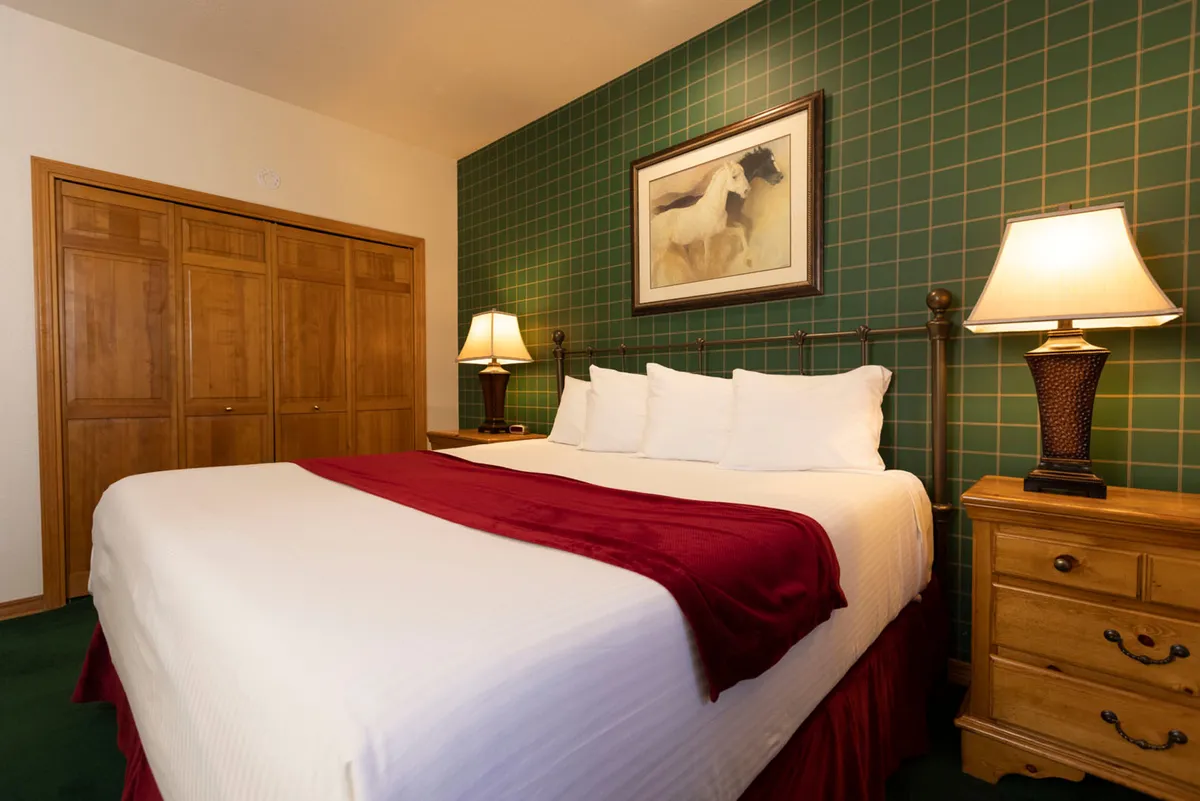 This image presents a bedroom with a bed dressed in a red throw blanket, a distinctive green checkered feature wall, wooden wardrobe, framed equestrian artwork, and softly glowing bedside lamps. The decor is welcoming and intimate, promoting a serene, restful retreat.