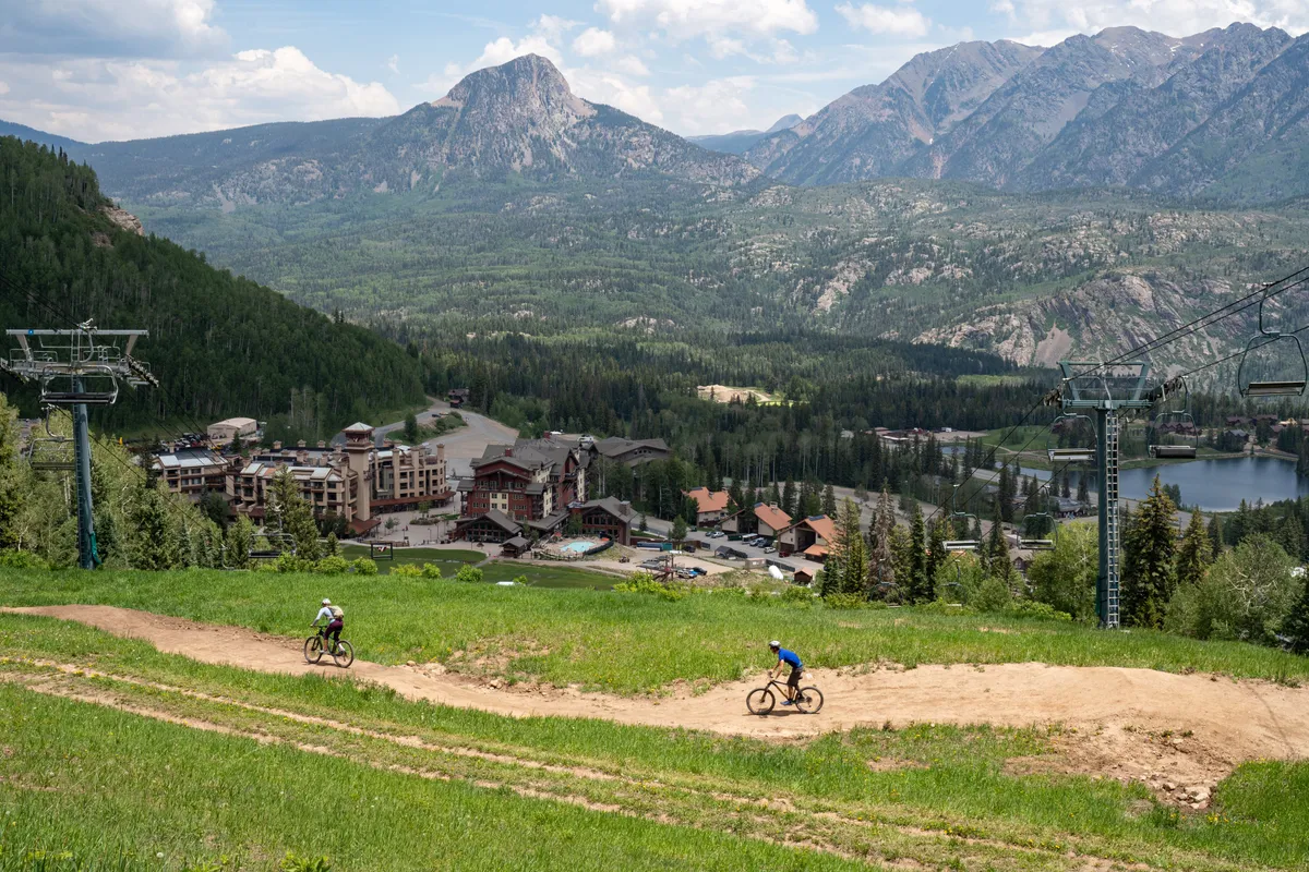 This elevated view showcases breathtaking mountains, a scenic biking trail, and Cascade Village’s resort buildings adjacent to a tranquil lake. The ski lift in the background hints at year-round recreation, while the open sky and expansive landscape evoke a sense of spaciousness and rejuvenation.